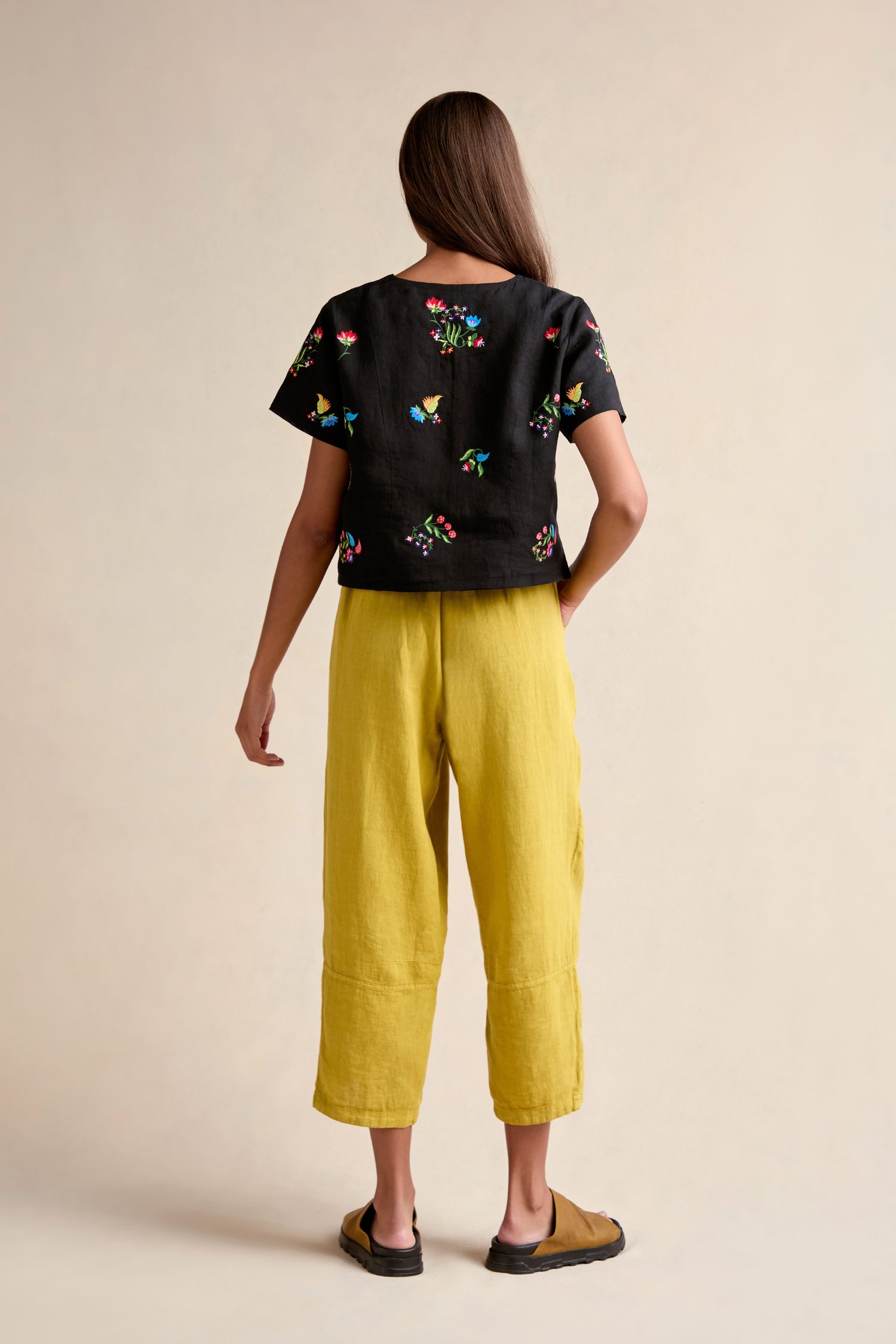 A person stands facing away, wearing the Vintage Floral Embroidered Jacket—a black short-sleeved top with colorful embroidery and a boxy silhouette—paired with yellow wide-leg pants and tan sandals against a neutral background.