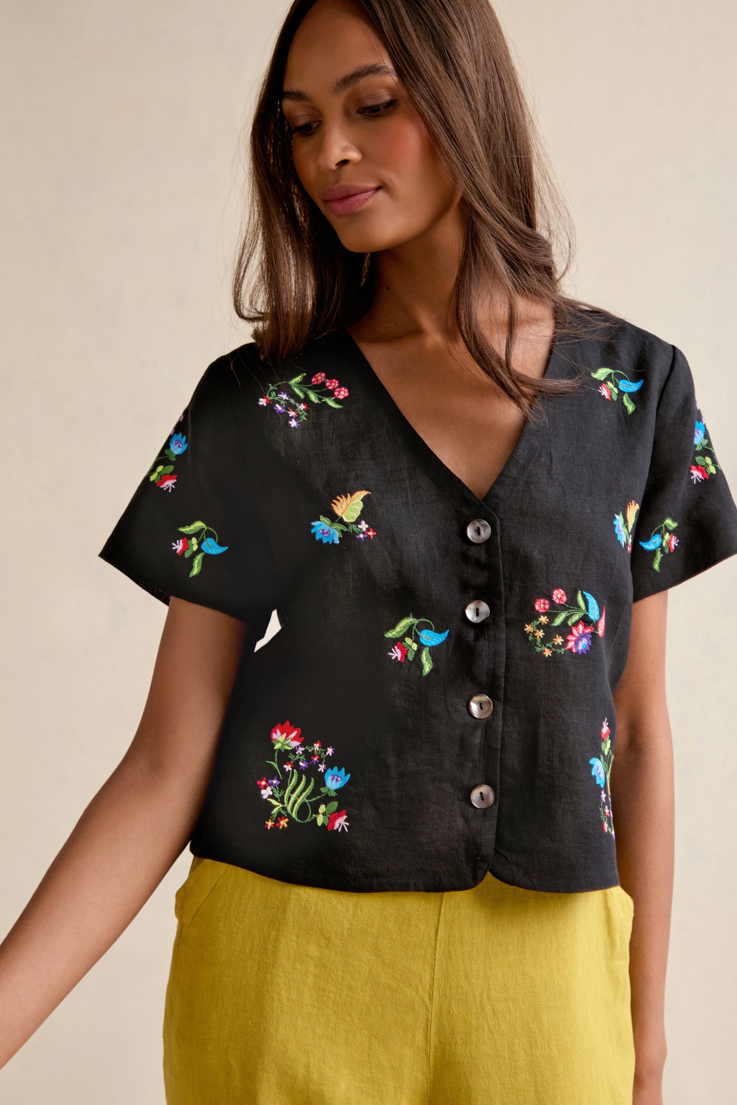 Woman wearing the Vintage Floral Embroidered Jacket—a black, short-sleeve button-up top with colorful floral embroidery—paired with yellow pants and styled in a boxy silhouette, stands against a plain background.