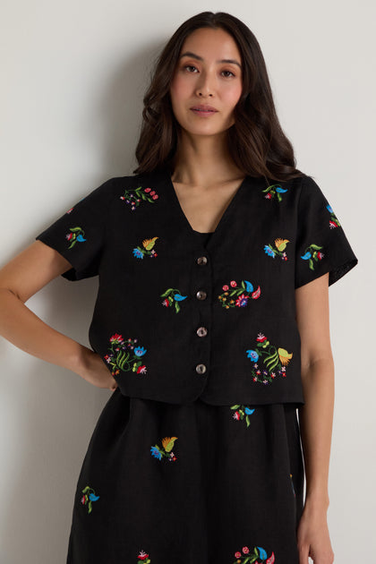 A woman stands against a plain wall wearing the Vintage Floral Embroidered Jacket, a black short-sleeve buttoned top with colorful bird and floral embroidery and a boxy silhouette for a modern touch.