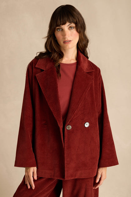 A woman stands against a plain beige background wearing the Conker Jumbo Cord Caban Jacket, styled as part of a loose-fitting burgundy broad wale cord suit.