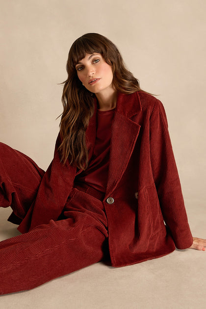 A woman sits on the floor against a neutral background, wearing the Conker Jumbo Cord Caban Jacket in burgundy with a coordinating top.