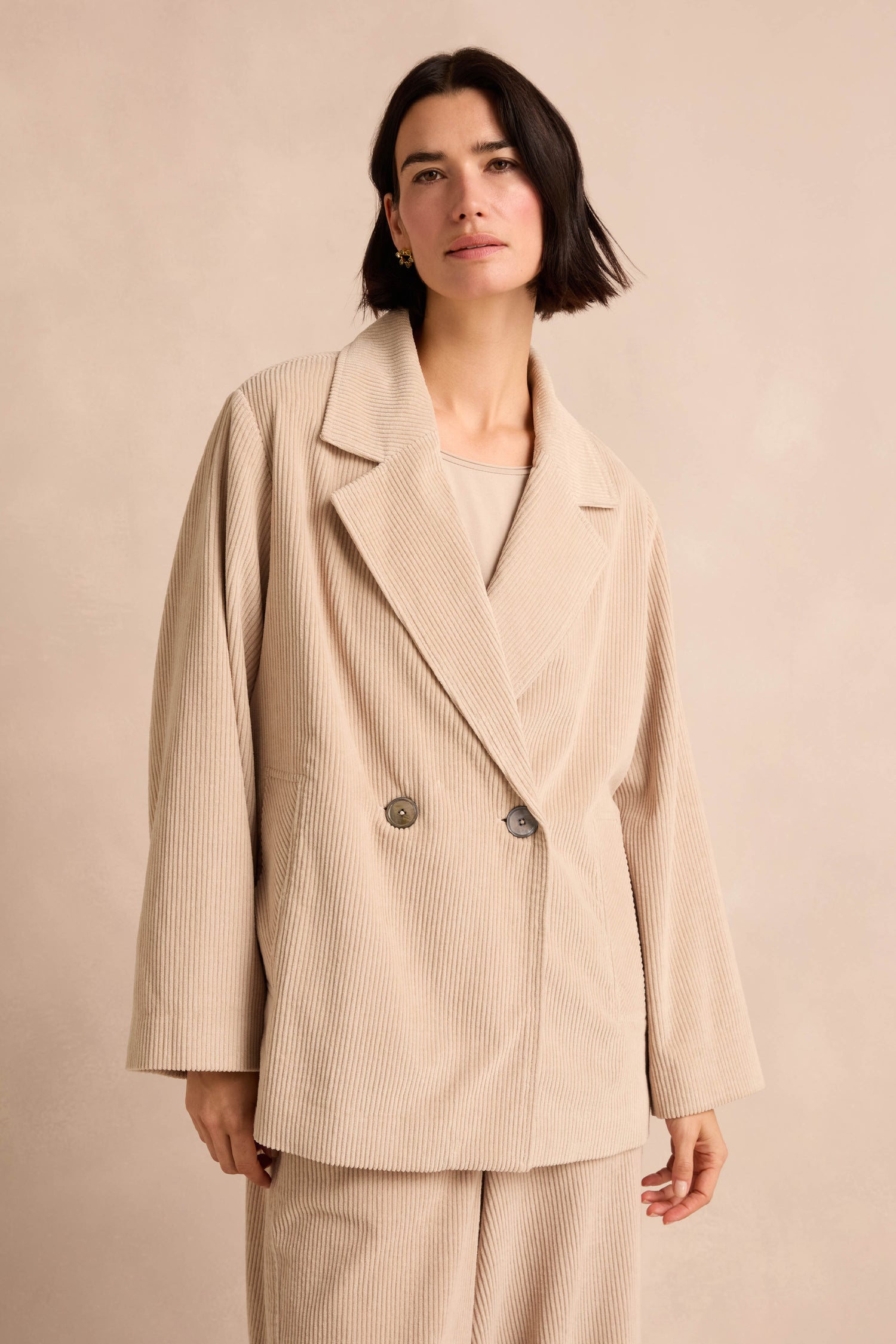 A woman with short dark hair wears the Ecru Jumbo Cord Caban Jacket, featuring pure cotton fabric and two buttons, along with matching pants, as she stands against a light neutral background.