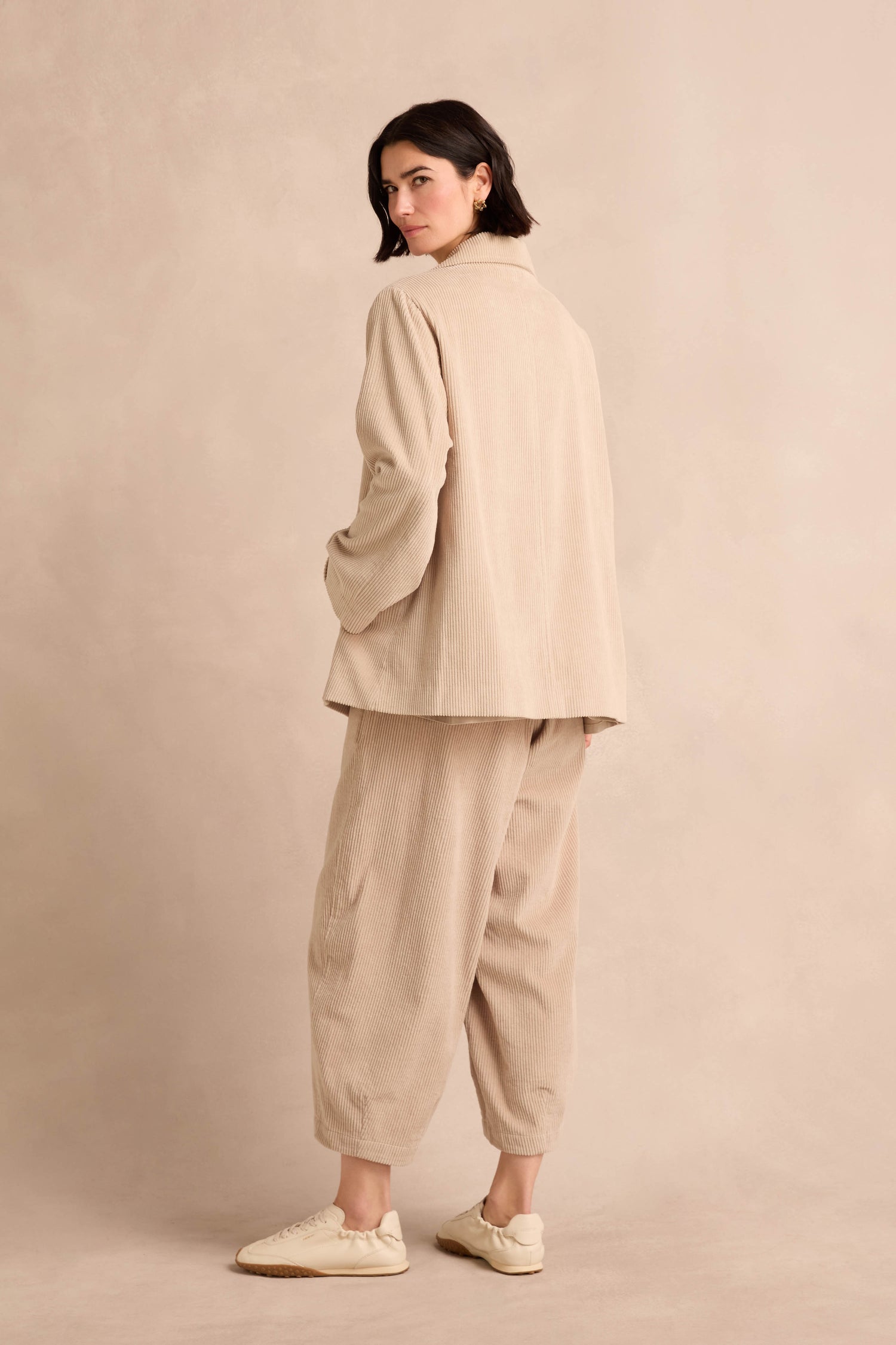 Person standing and looking over their shoulder, wearing an Ecru Jumbo Cord Caban Jacket and white sneakers, against a plain beige background.