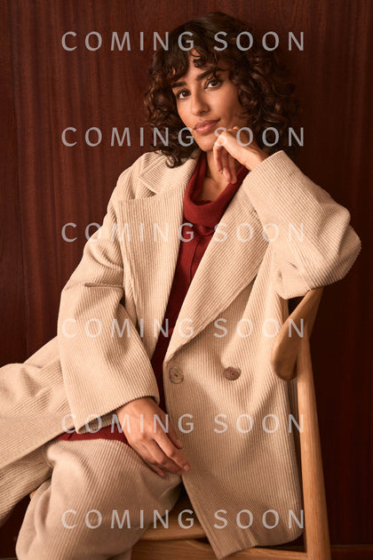 A person wearing the Ecru Jumbo Cord Caban Jacket sits on a wooden chair against a brown background, with the words “COMING SOON” repeated over the image.