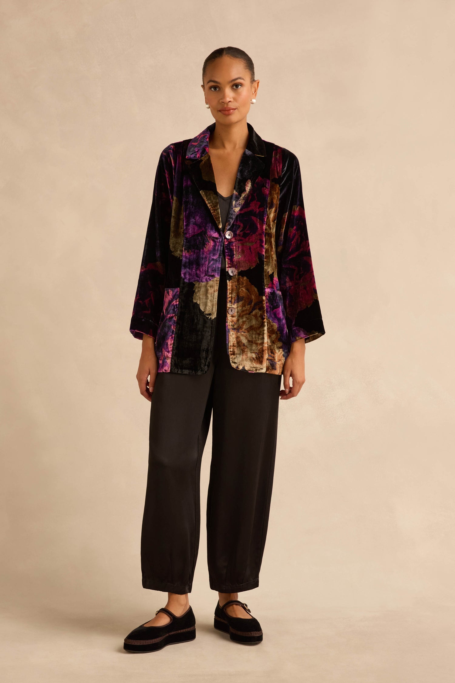 A person wearing a Midnight Peonies Velvet Relaxed Blazer in black, purple, and gold floral velvet, with black trousers and black Mary Jane shoes.