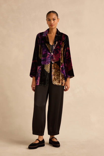 A person wearing a Midnight Peonies Velvet Relaxed Blazer in black, purple, and gold floral velvet, with black trousers and black Mary Jane shoes.