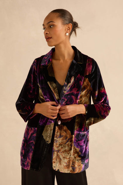 A woman wears the Midnight Peonies Velvet Relaxed Blazer, a relaxed-fit, button-front velvet jacket featuring a large, dark-toned floral print in shades of purple and gold.