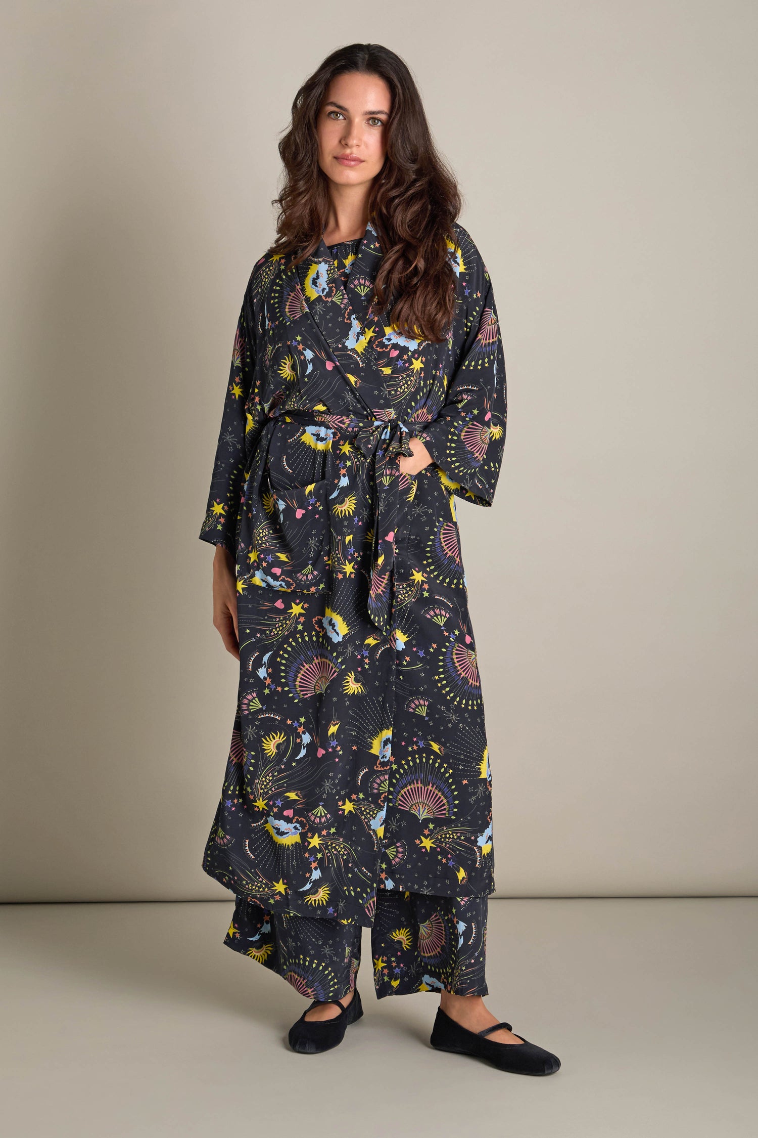 A person is wearing the exquisite Celestial Dreams Robe, characterized by its long length and dark fabric adorned with vibrant, colorful patterns, as they stand against a plain background.