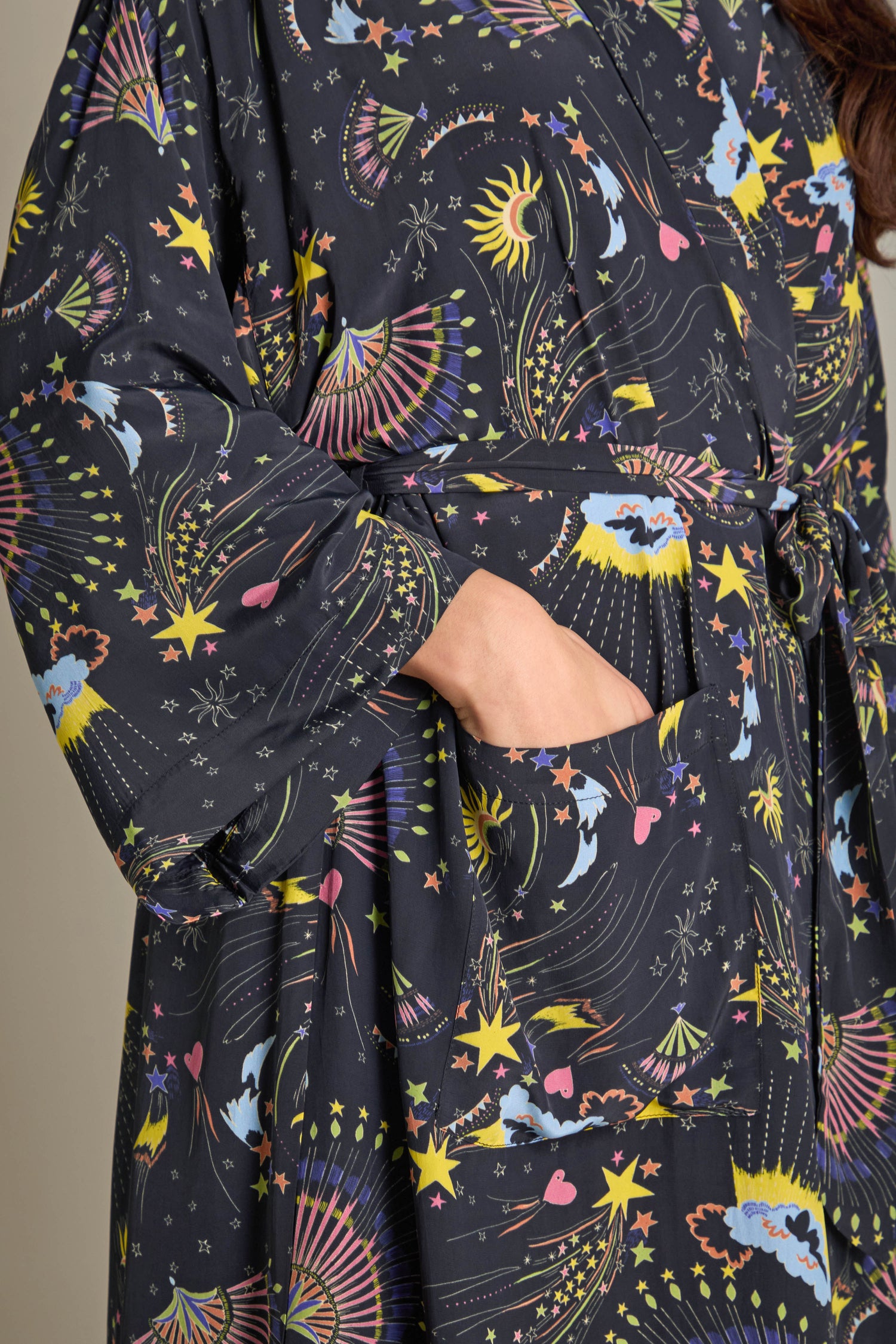 A person wearing the Celestial Dreams Robe, crafted from dark, silky fabric and featuring vibrant celestial designs, exuding luxurious comfort while casually resting a hand in one of its pockets.