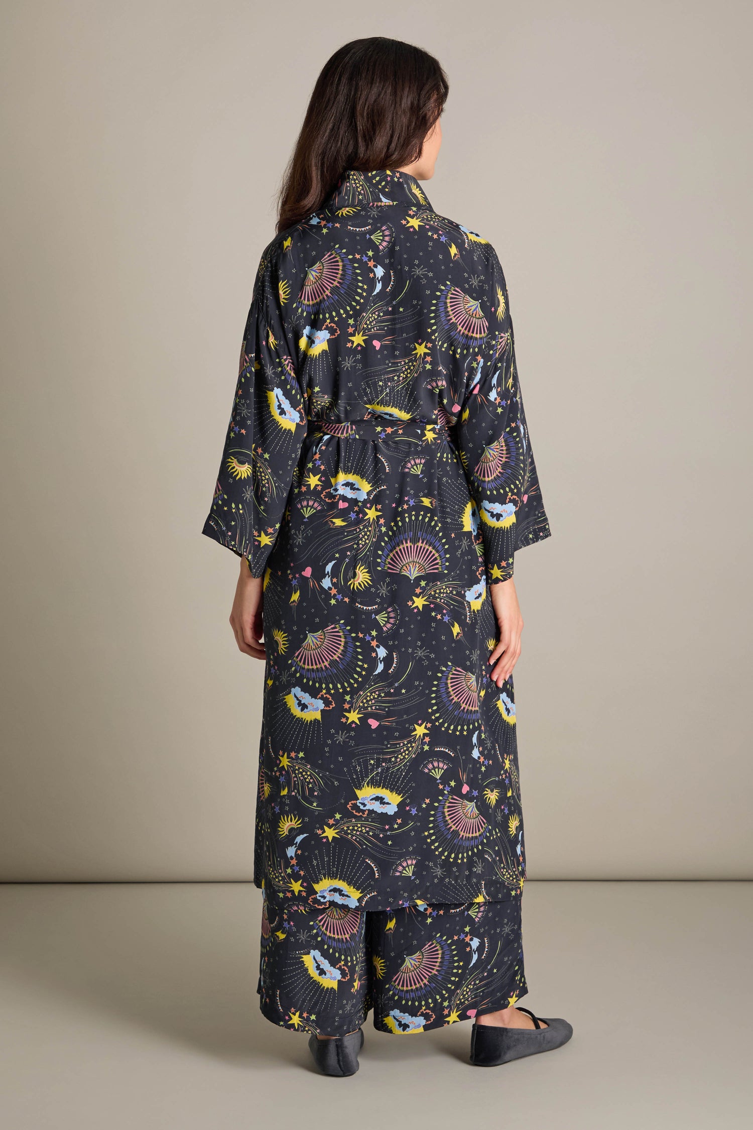 Draped in the Celestial Dreams Robe, featuring colorful abstract designs on silky black fabric, a person stands with their back to the camera, embodying luxurious comfort and elegance in the simplicity of a plain room.