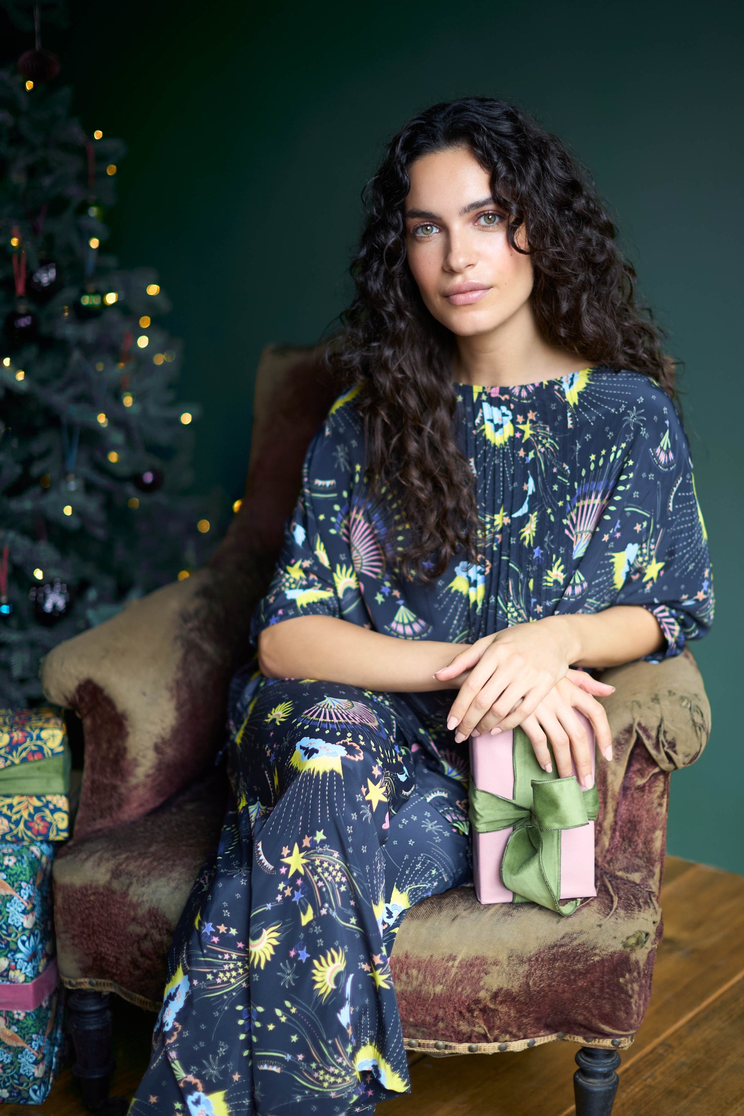 Apologies for the confusion. Here's a revised description with keywords integrated: A joyful woman with curly hair sits on a chair, wearing the Celestial Dreams Pyjama Set, and cradling a gift box adorned with a green ribbon beside a beautifully decorated Christmas tree—perfect for capturing the essence of festive cheer.