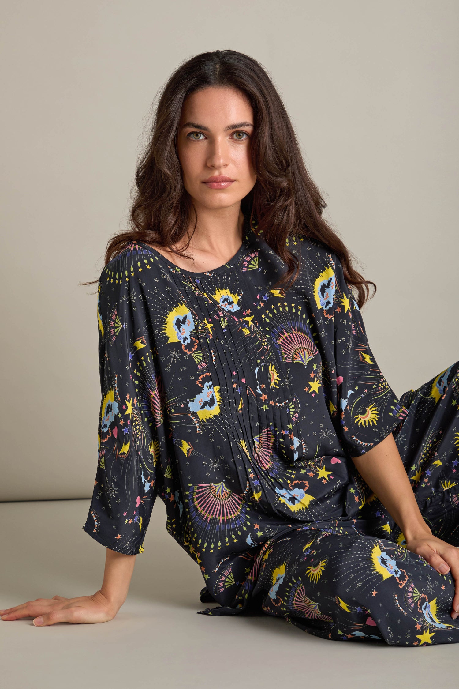 A person with long hair wearing the Celestial Dreams Pyjama Set sits on the floor against a plain backdrop, embodying a luxurious night's sleep.