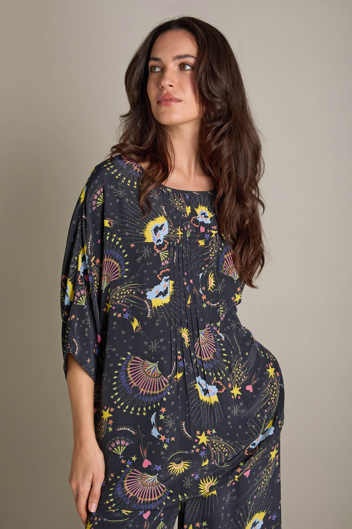 A woman in a vibrant Celestial Dreams Pyjama Set stands against a neutral backdrop, gazing to the side, evoking the essence of a celestial dreamscape.