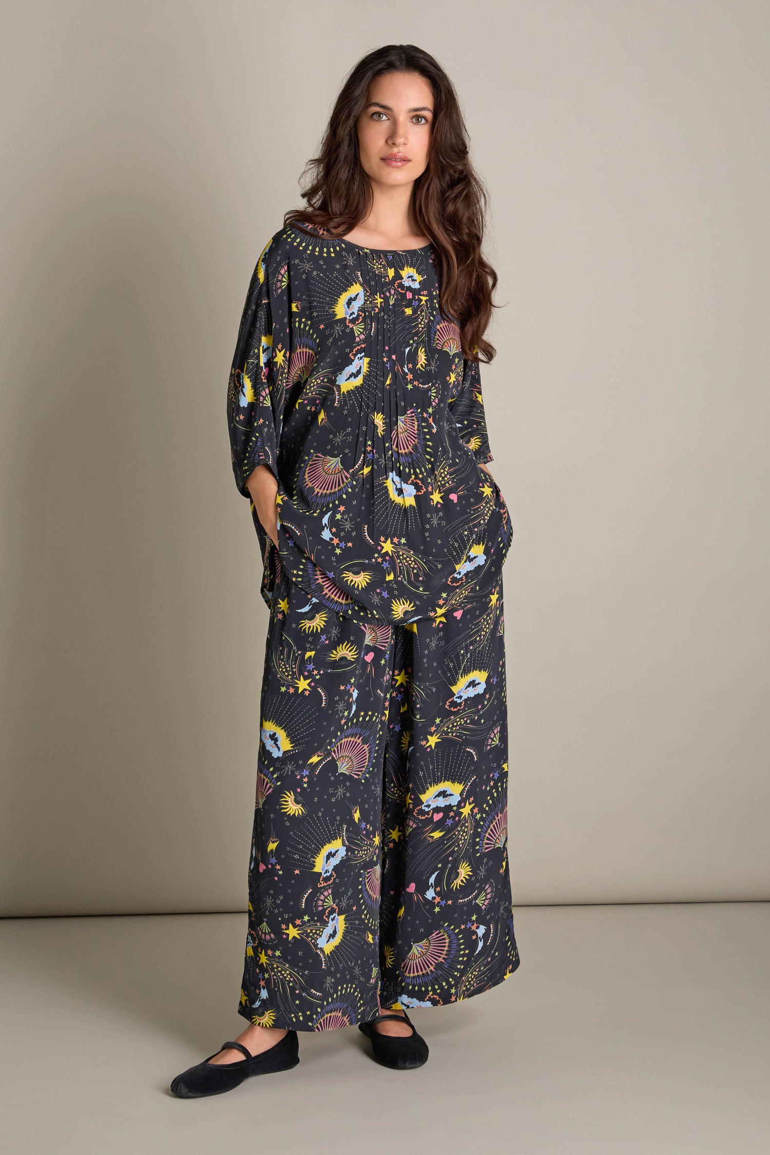 A woman stands against a plain background, wearing the Celestial Dreams Pyjama Set—featuring loose-fitting, dark fabric adorned with abstract designs—and exudes an enchanting allure, as her hands rest casually in the pockets.