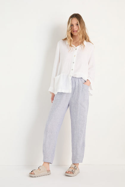A person stands against a white background wearing a loose white button-up top, beige sandals, and Ticking Linen Stripe Bubble Trousers with an elasticated waist.