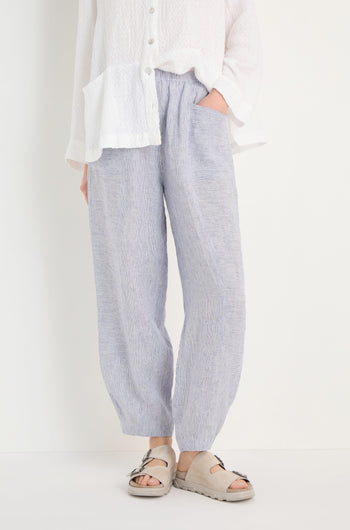 Person wearing Ticking Linen Stripe Bubble Trousers with an elasticated waist, a white textured button-up shirt, and beige sandals, standing against a plain white background with hands in the pants' pockets.