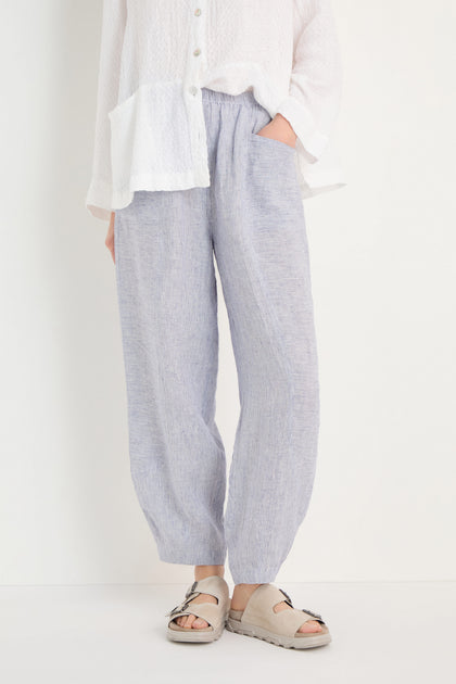 Person wearing Ticking Linen Stripe Bubble Trousers with an elasticated waist, a white textured button-up shirt, and beige sandals, standing against a plain white background with hands in the pants' pockets.