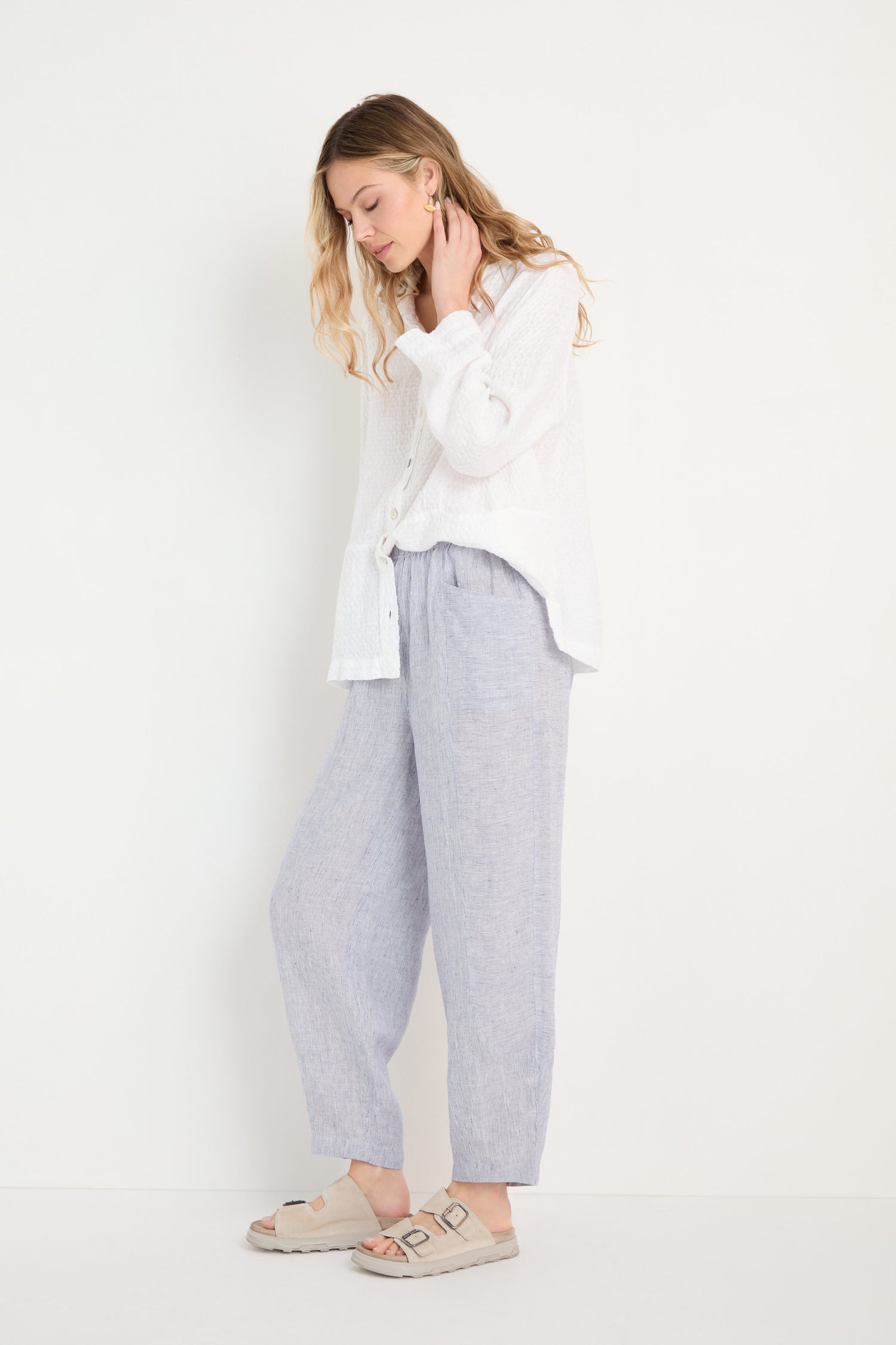 A woman stands against a white wall wearing a white button-up shirt, Ticking Linen Stripe Bubble Trousers in light gray with an elasticated waist, and beige sandals.