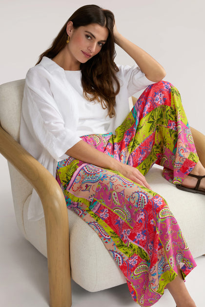 A woman sits on a cushioned chair, wearing a white top and brightly colored Tropical Patchwork Wide Trouser.