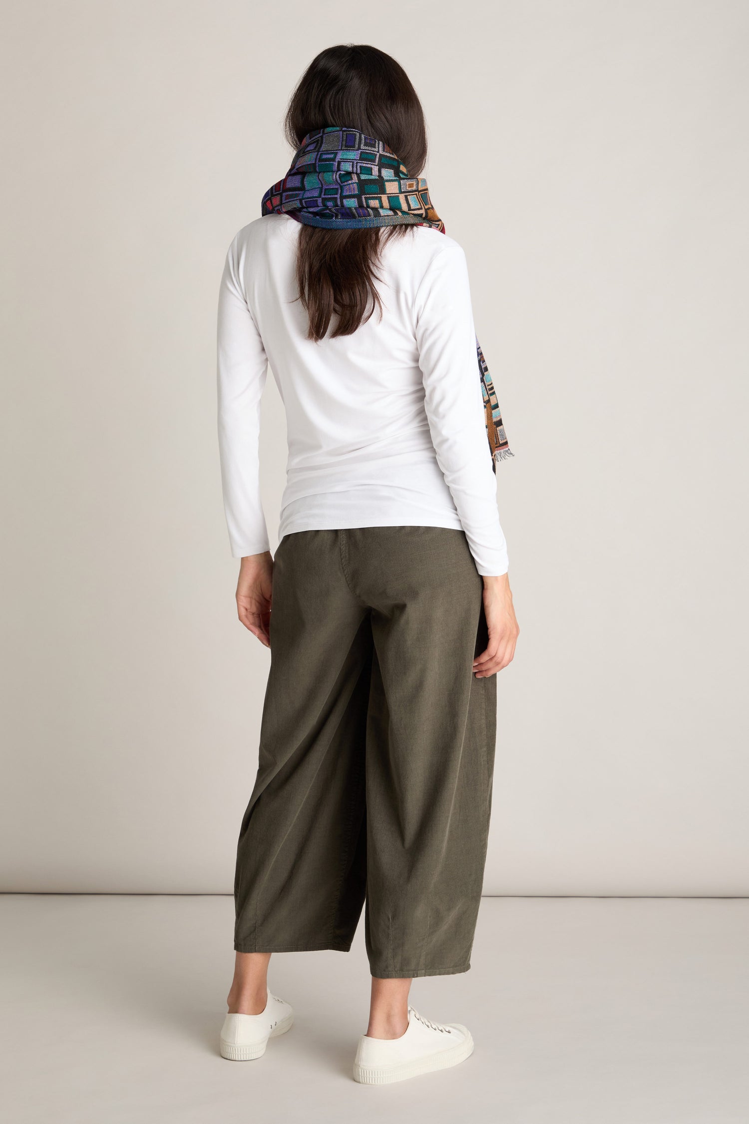 A person stands with their back facing the camera, wearing a white long-sleeve shirt, green Pinwale Cord Bubble Trousers with an elasticated waist, a colorful scarf, and white sneakers.