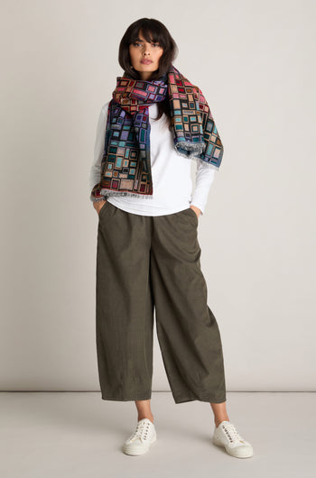 A person standing against a plain background, wearing a white long-sleeve shirt, olive green Pinwale Cord Bubble Trousers, white sneakers, and a colorful geometric-patterned scarf.