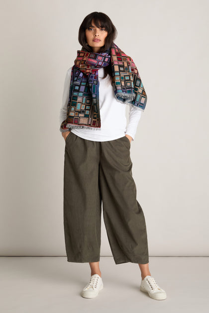 A person standing against a plain background, wearing a white long-sleeve shirt, olive green Pinwale Cord Bubble Trousers, white sneakers, and a colorful geometric-patterned scarf.