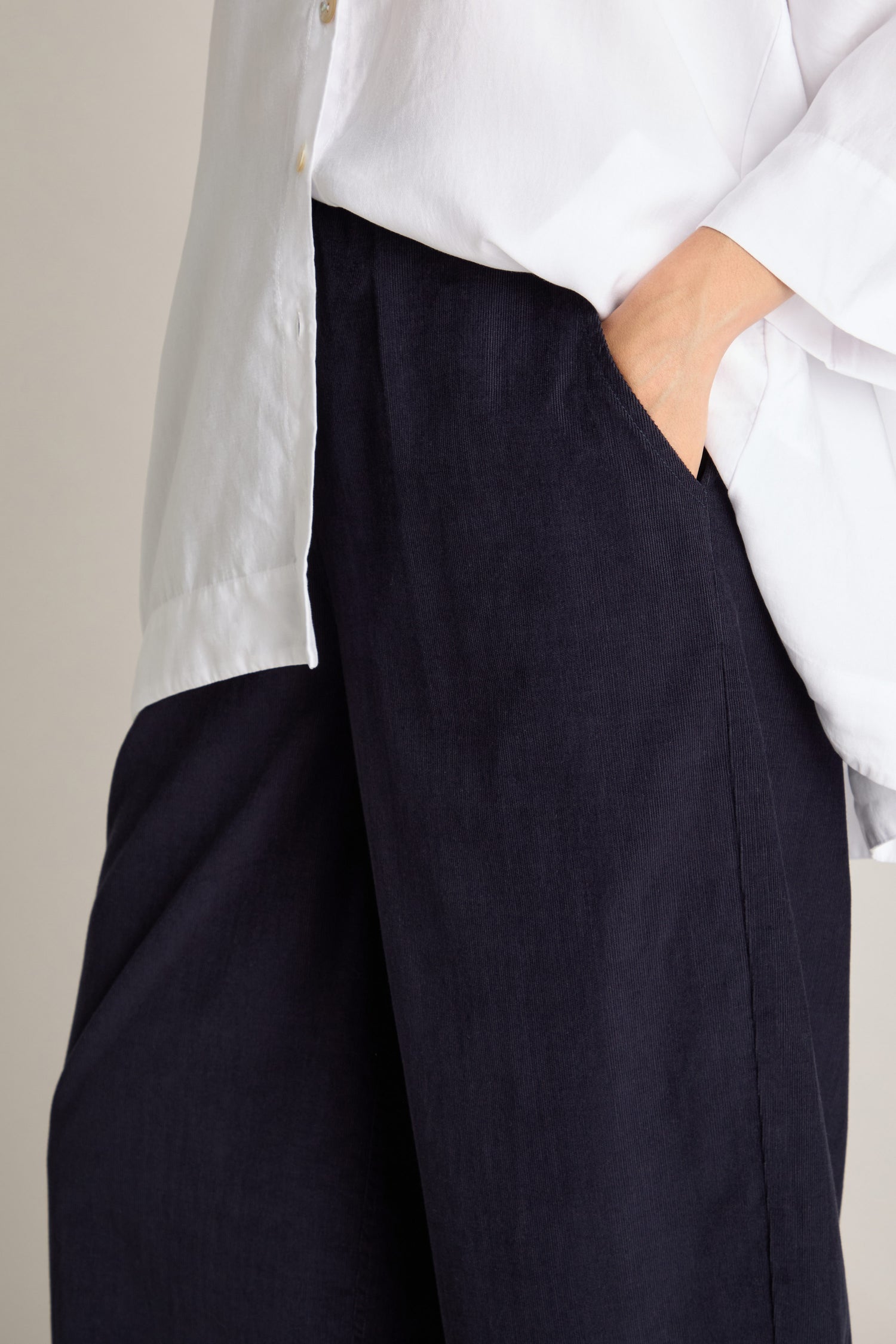 A person wearing a white button-up shirt and dark Pinwale Cord Bubble Trousers with an elasticated waist, casually placing their right hand in the trouser pocket.