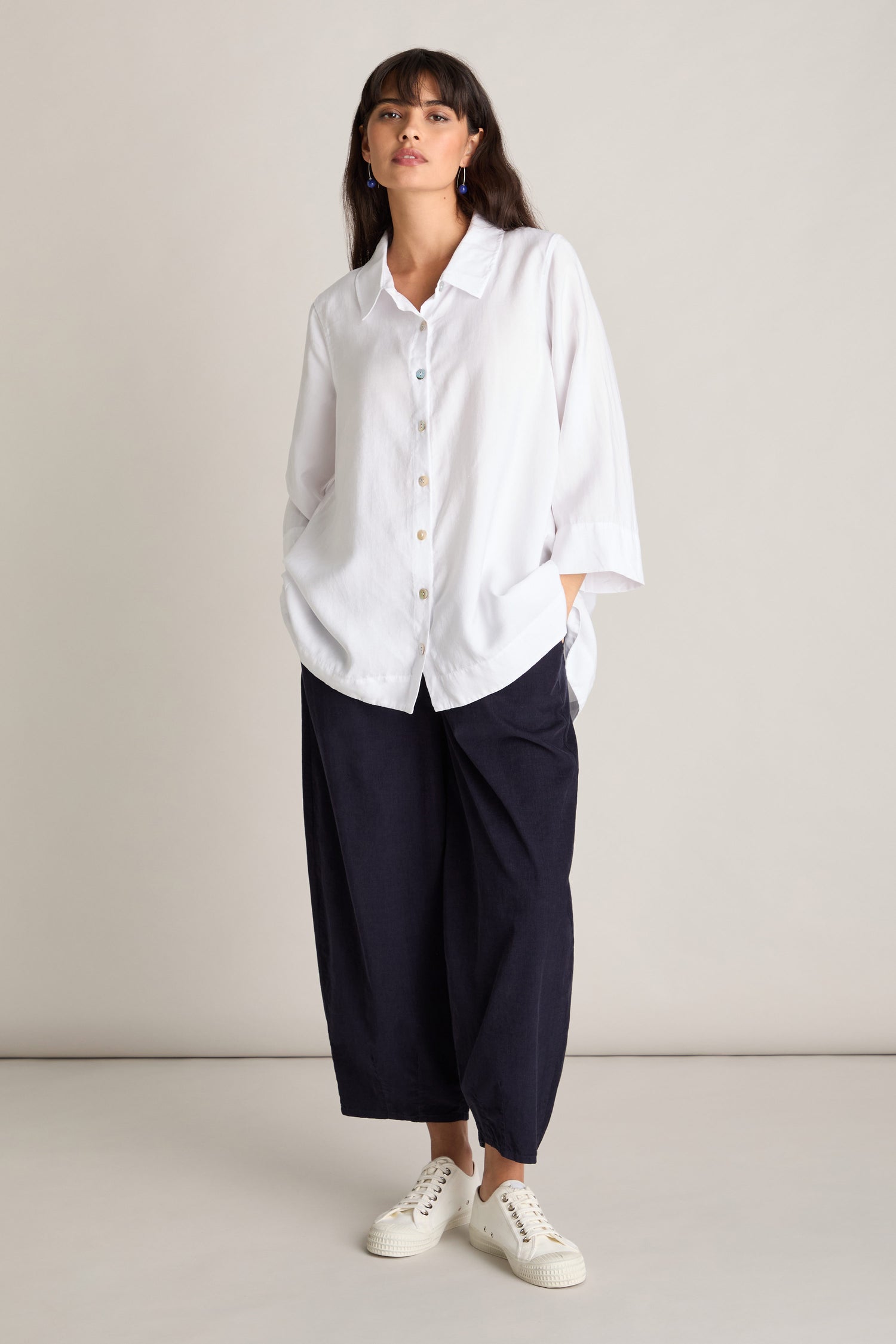 A person stands against a light background, wearing a loose white button-up shirt, dark Pinwale Cord Bubble Trousers with an elasticated waist, and white sneakers.