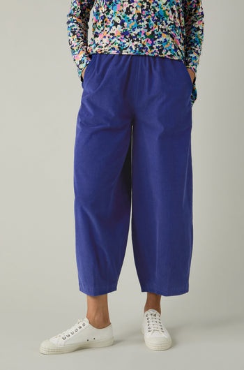 A person wearing cropped-length Pinwale Cord Bubble Trousers in blue, a colorful long-sleeve top, and white sneakers stands with hands in pockets against a plain background.