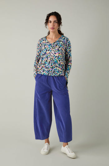 A person stands against a plain background wearing a colorful floral top, white sneakers, and the Pinwale Cord Bubble Trouser in cropped blue style, posing with hands in pockets.
