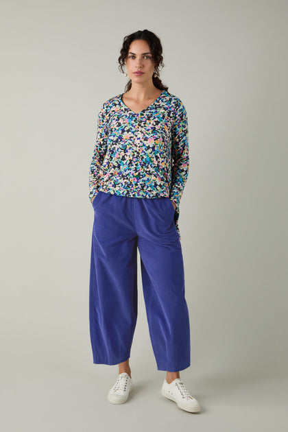 A person stands against a plain background wearing a colorful floral top, white sneakers, and the Pinwale Cord Bubble Trouser in cropped blue style, posing with hands in pockets.