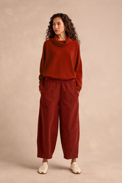 Standing against a beige background, the person wears a rust-colored long-sleeve top paired with the Conker Pinwale Cord Bubble Trouser—a relaxed fit style in fine pincord—alongside white sneakers, with their hands in their pockets.