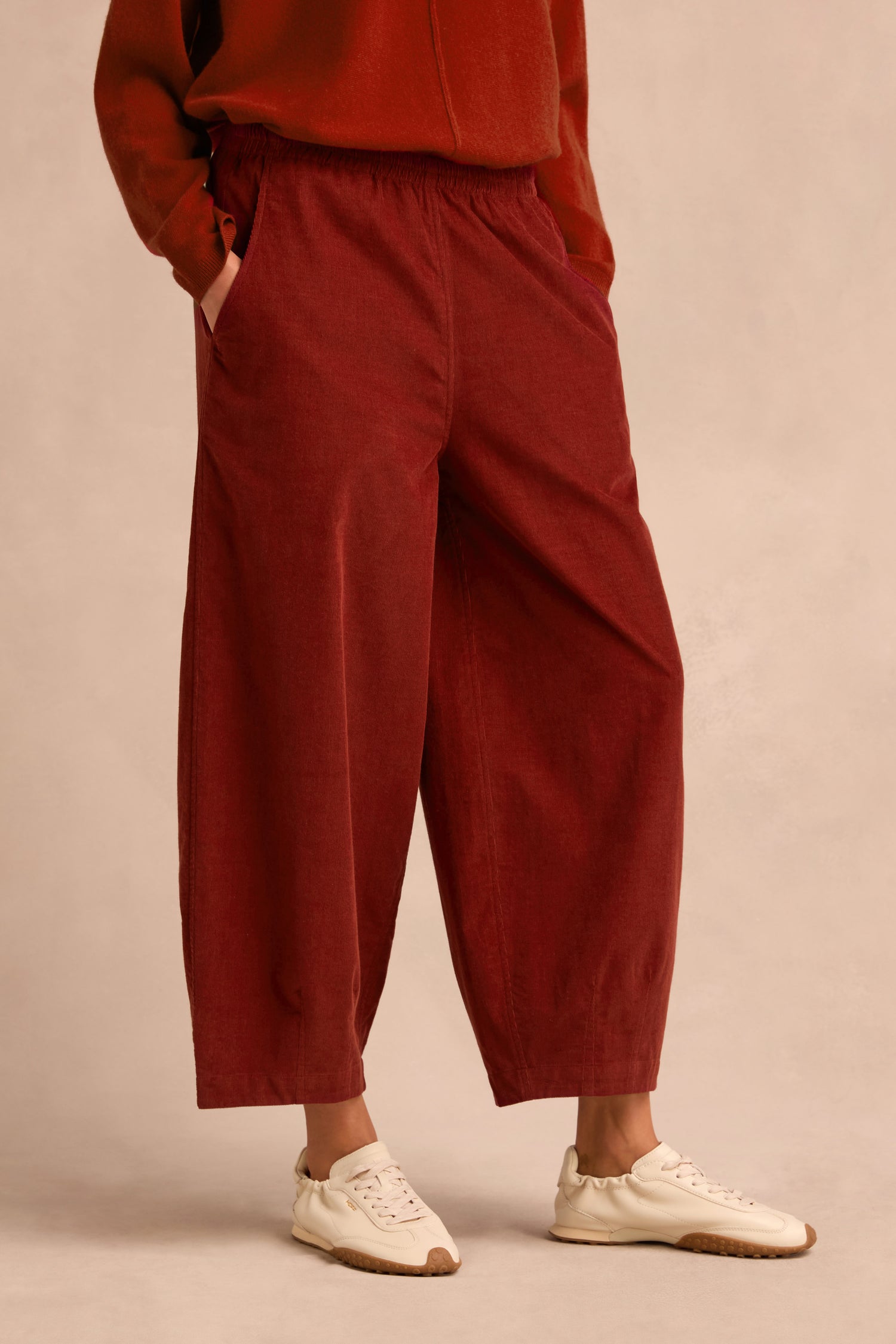 A person stands with hands in pockets against a plain beige background, wearing the Conker Pinwale Cord Bubble Trouser in red with a relaxed fit, paired with a matching red top and white sneakers.