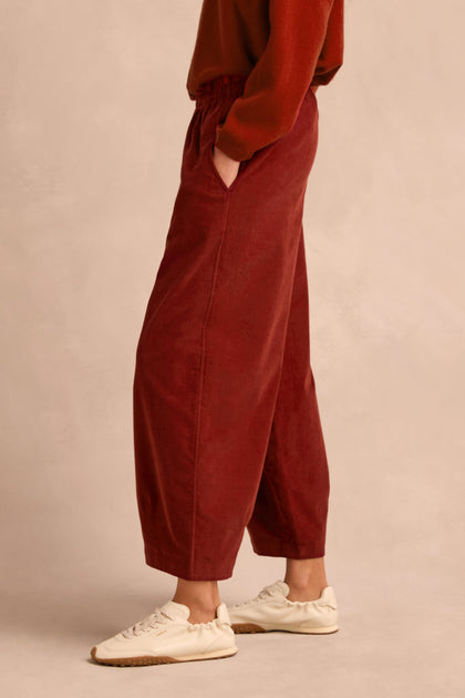 A person wearing the Conker Pinwale Cord Bubble Trouser, styled with a red sweater and white sneakers, stands sideways with hands in pockets against a neutral background.