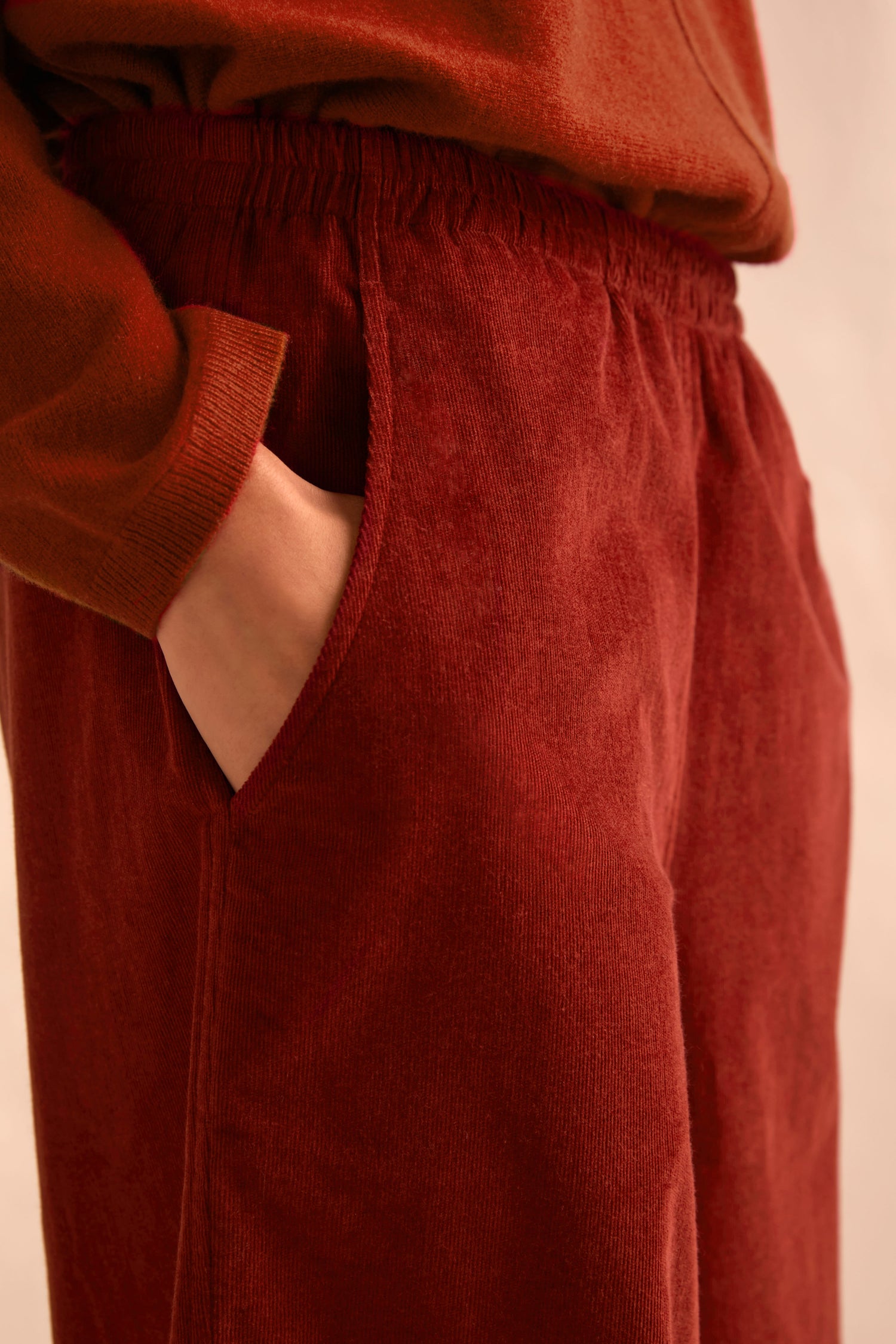 A person wears the Conker Pinwale Cord Bubble Trouser in rust—a relaxed-fit fine pincord corduroy pant with an elastic waistband—paired with a matching sweater, one hand casually tucked into a pocket.
