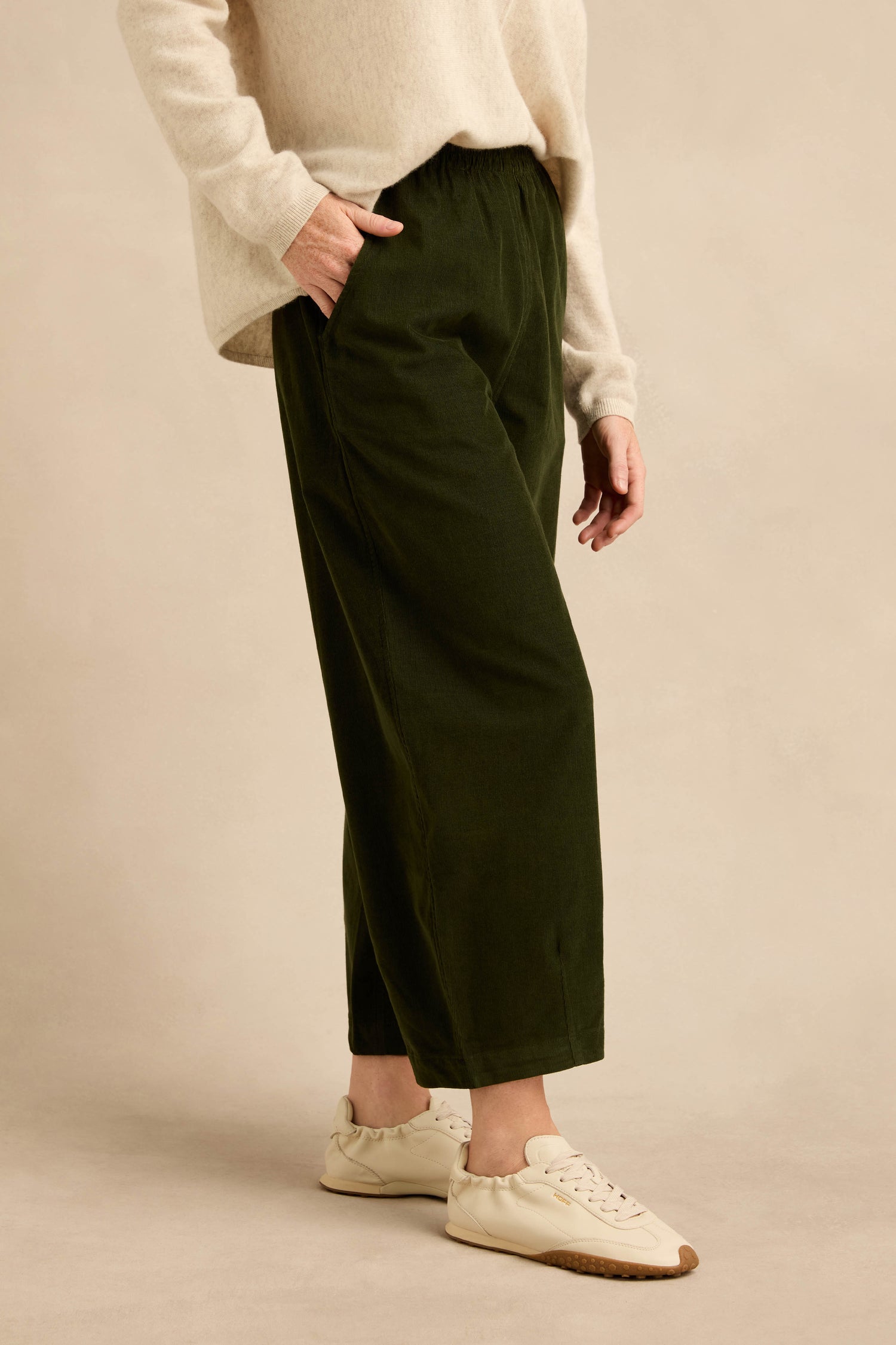 A person wearing a cream sweater, Forest Pinwale Cord Bubble Trousers in dark green, and cream sneakers stands with one hand in their pocket against a beige background.