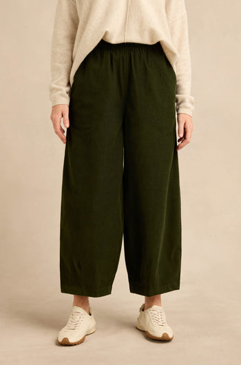 Person wearing a beige sweater, Forest Pinwale Cord Bubble Trousers in a relaxed wide-leg fit, and white sneakers, standing against a plain light background.