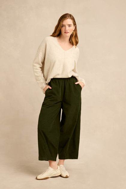 A woman stands against a plain background, hands in her pockets, wearing a beige V-neck sweater and the Forest Pinwale Cord Bubble Trousers in dark green with a relaxed fit, paired with white sneakers.