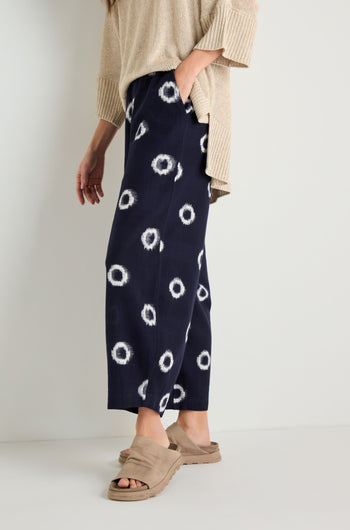 A person stands against a plain background wearing a beige sweater, Ikat Circles Cotton Bubble Trousers in indigo ecru with a white circular design, and beige sandals.
