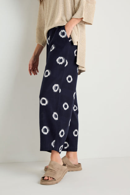 A person stands against a plain background wearing a beige sweater, Ikat Circles Cotton Bubble Trousers in indigo ecru with a white circular design, and beige sandals.