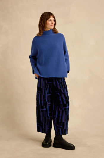 A person stands against a plain background, hands in pockets, wearing a loose blue sweater, black boots, and the Layered Geometric Cord Trousers—wide-leg pants featuring a bold geometric pattern—while looking off to the side.