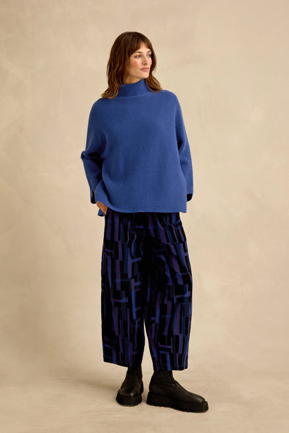 A person stands against a plain background, hands in pockets, wearing a loose blue sweater, black boots, and the Layered Geometric Cord Trousers—wide-leg pants featuring a bold geometric pattern—while looking off to the side.