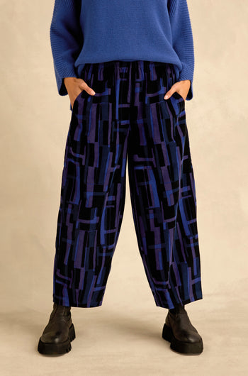 A person wearing the Layered Geometric Cord Trousers—featuring a striking black and blue geometric pattern—paired with a blue sweater and black boots, stands with hands in their pockets.
