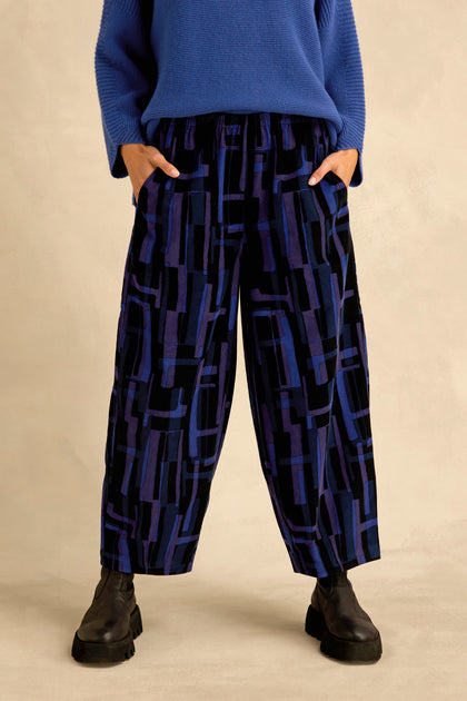 A person wearing the Layered Geometric Cord Trousers—featuring a striking black and blue geometric pattern—paired with a blue sweater and black boots, stands with hands in their pockets.