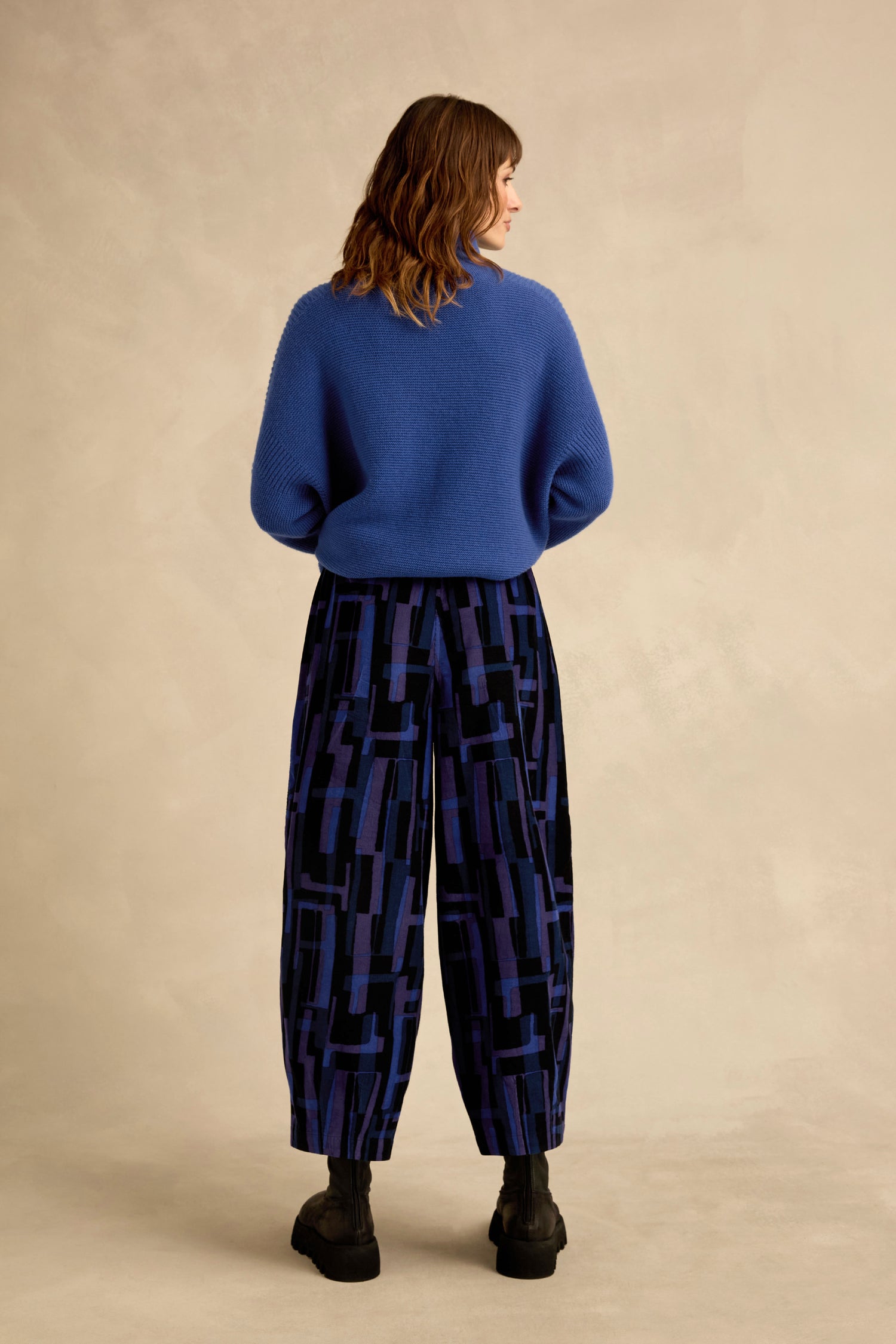 A woman stands with her back to the camera wearing a blue sweater, black boots, and Layered Geometric Cord Trousers against a neutral background.