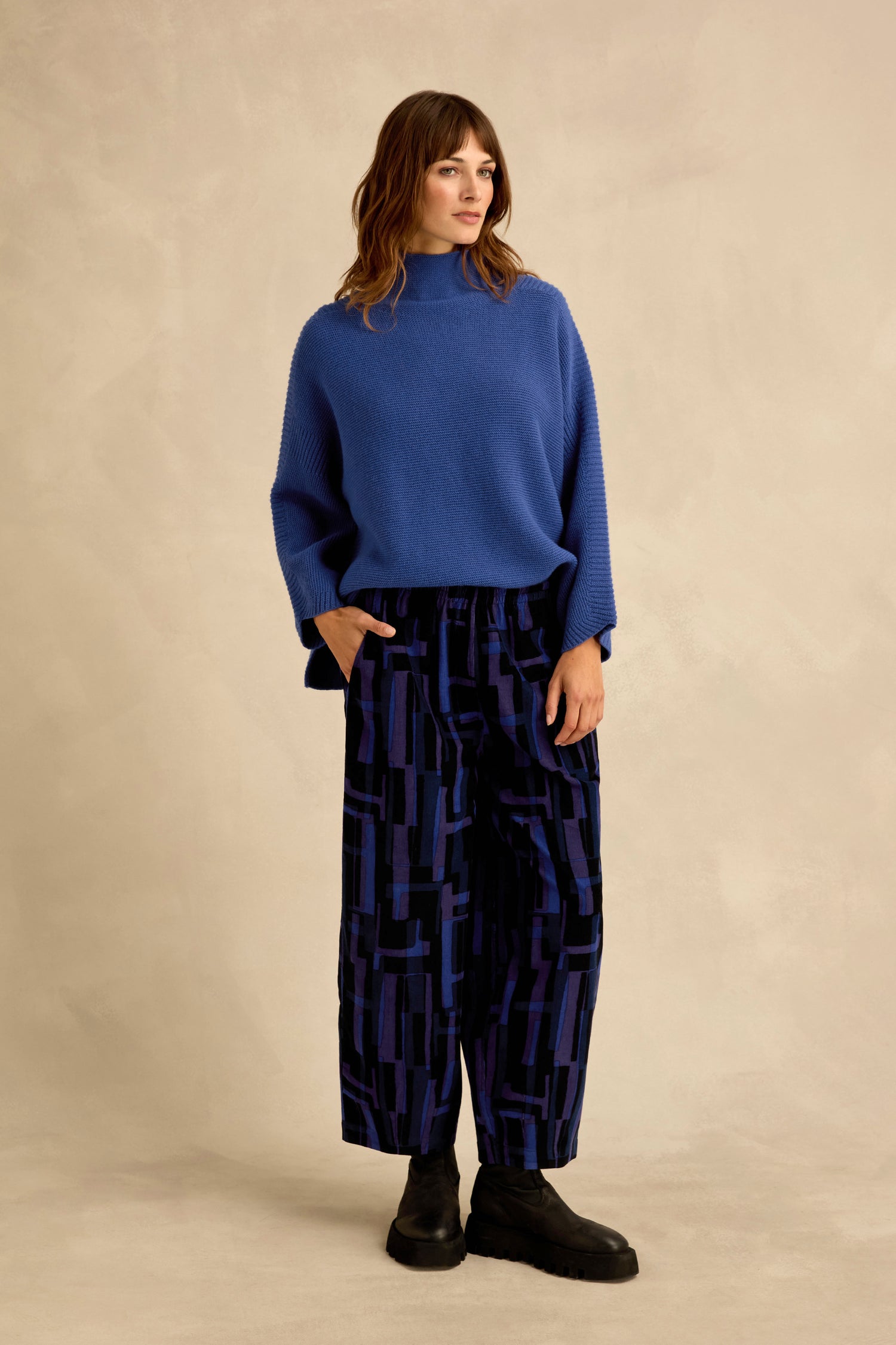 A woman stands against a beige background wearing a blue sweater, black shoes, and the Layered Geometric Cord Trousers featuring a black and blue geometric pattern and wide-leg silhouette, with her hands in her pockets.