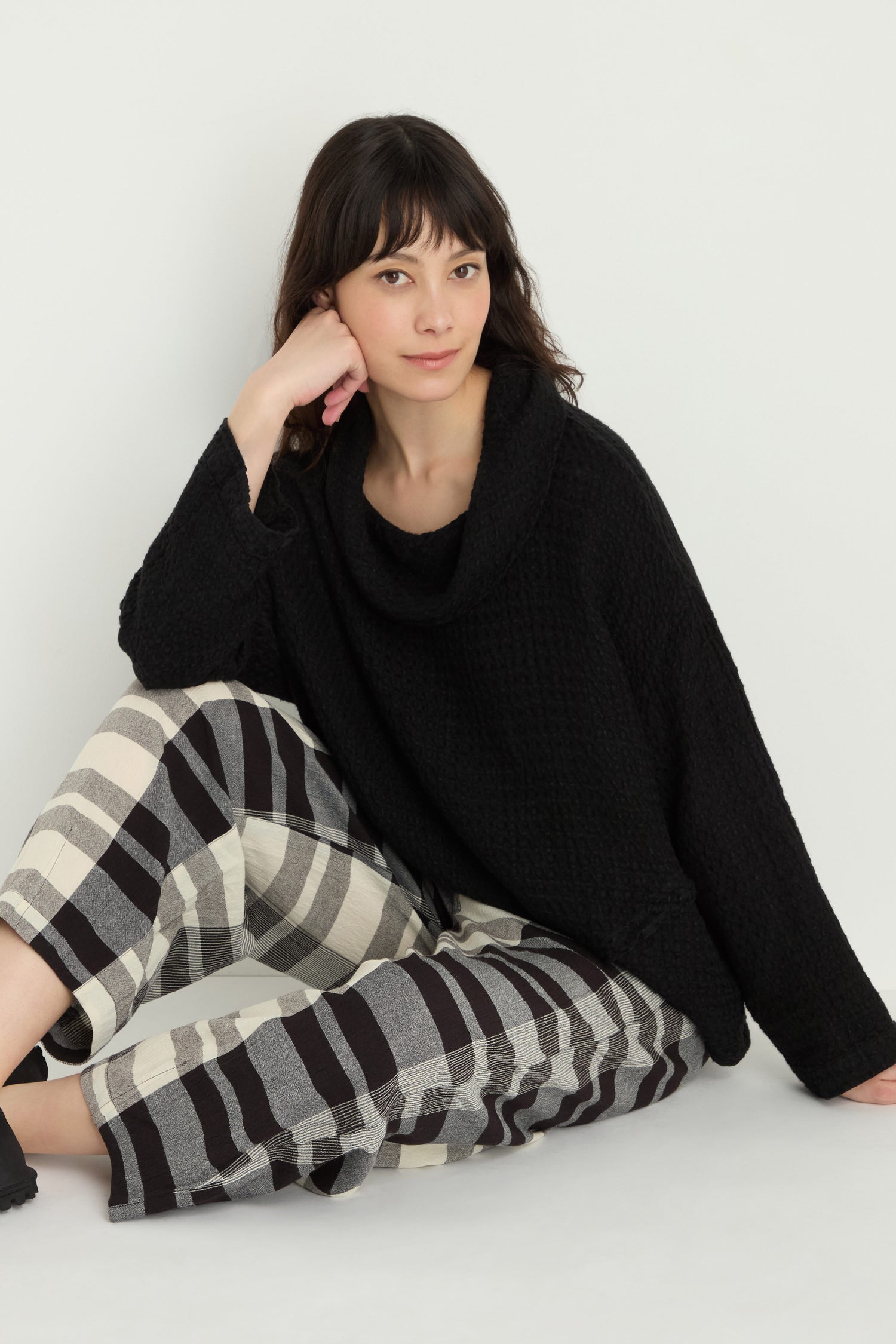 A woman sits on the floor against a white background, wearing a black textured sweater and Monochrome Crinkle Check Bubble Trouser with an elasticated waist in striped black, white, and gray.