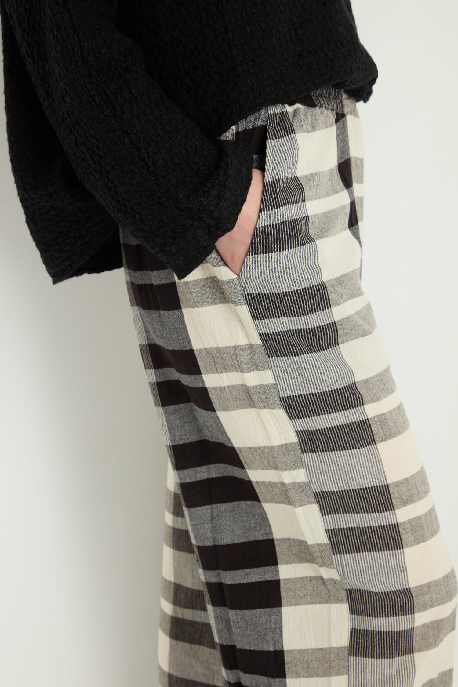 A person stands against a white background, wearing loose, high-waisted Monochrome Crinkle Check Bubble Trousers with one hand in the pocket and a black textured sweater.