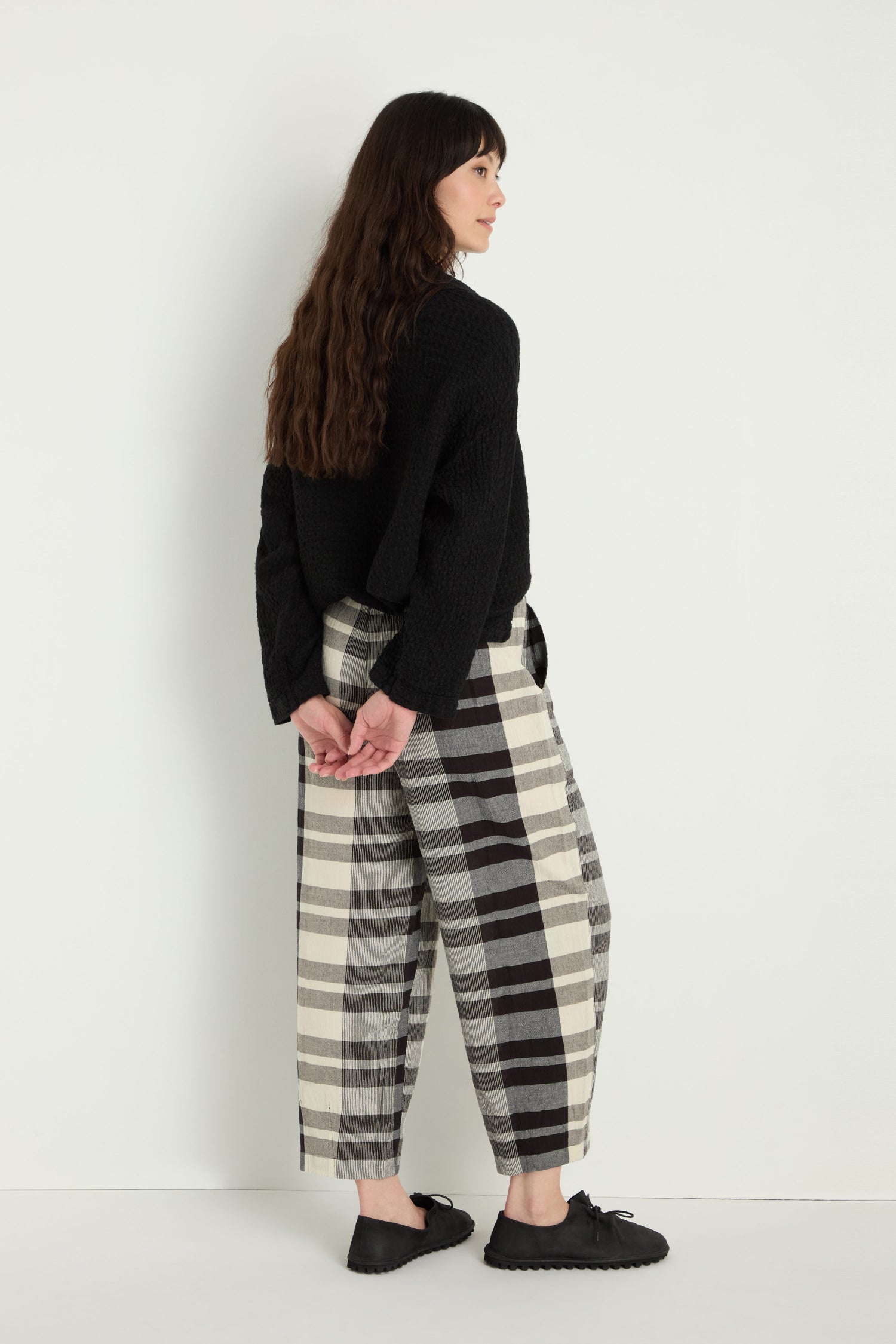 A person with long brown hair, wearing a black sweater, Monochrome Crinkle Check Bubble Trousers with an elasticated waist, and black shoes stands against a white wall with their hands clasped behind their back.