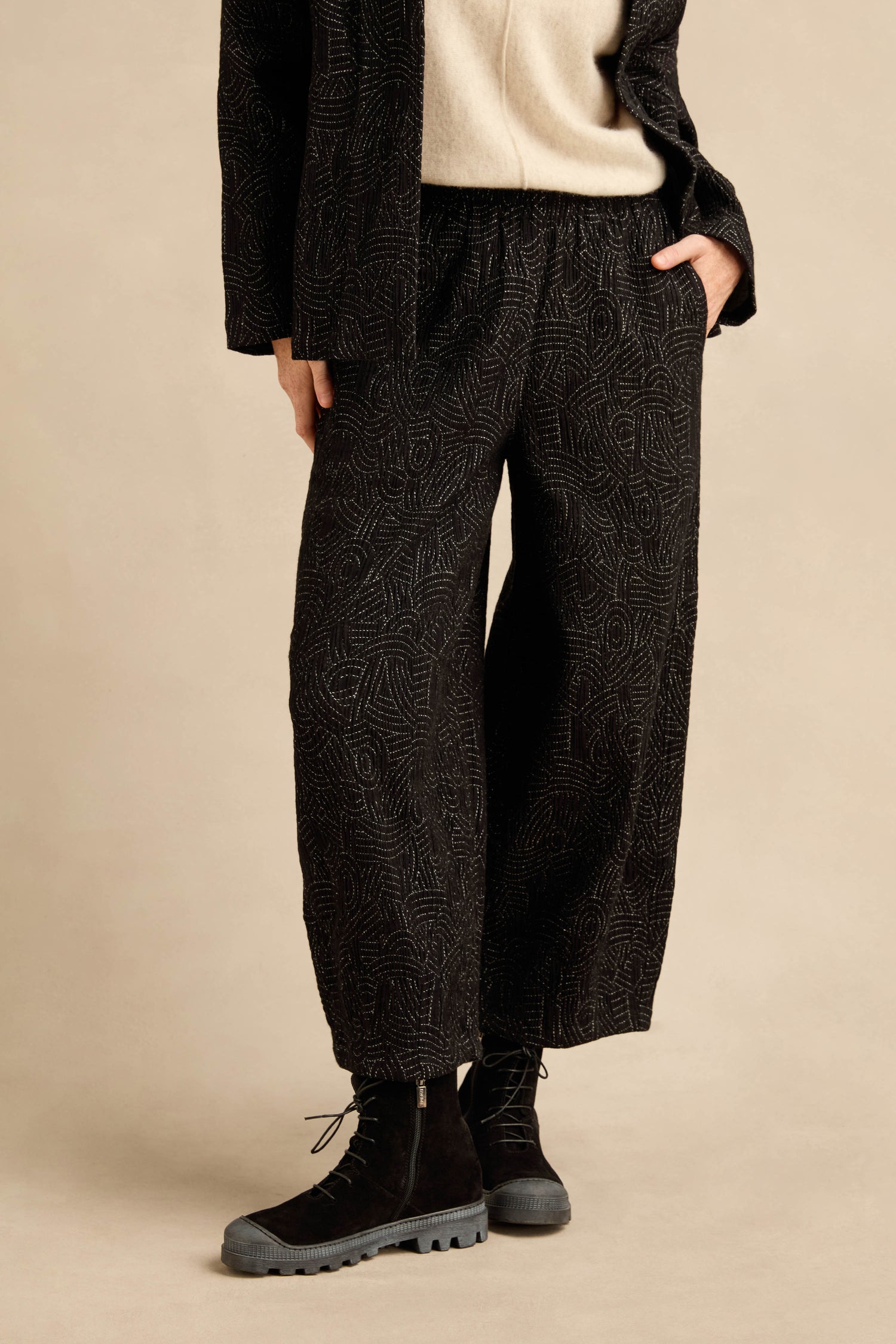 Person wearing the Stitched Jacquard Bubble Trouser in black with a cropped leg, paired with a cream top, a black jacket, and black lace-up boots, standing against a beige background. Their face is not visible.